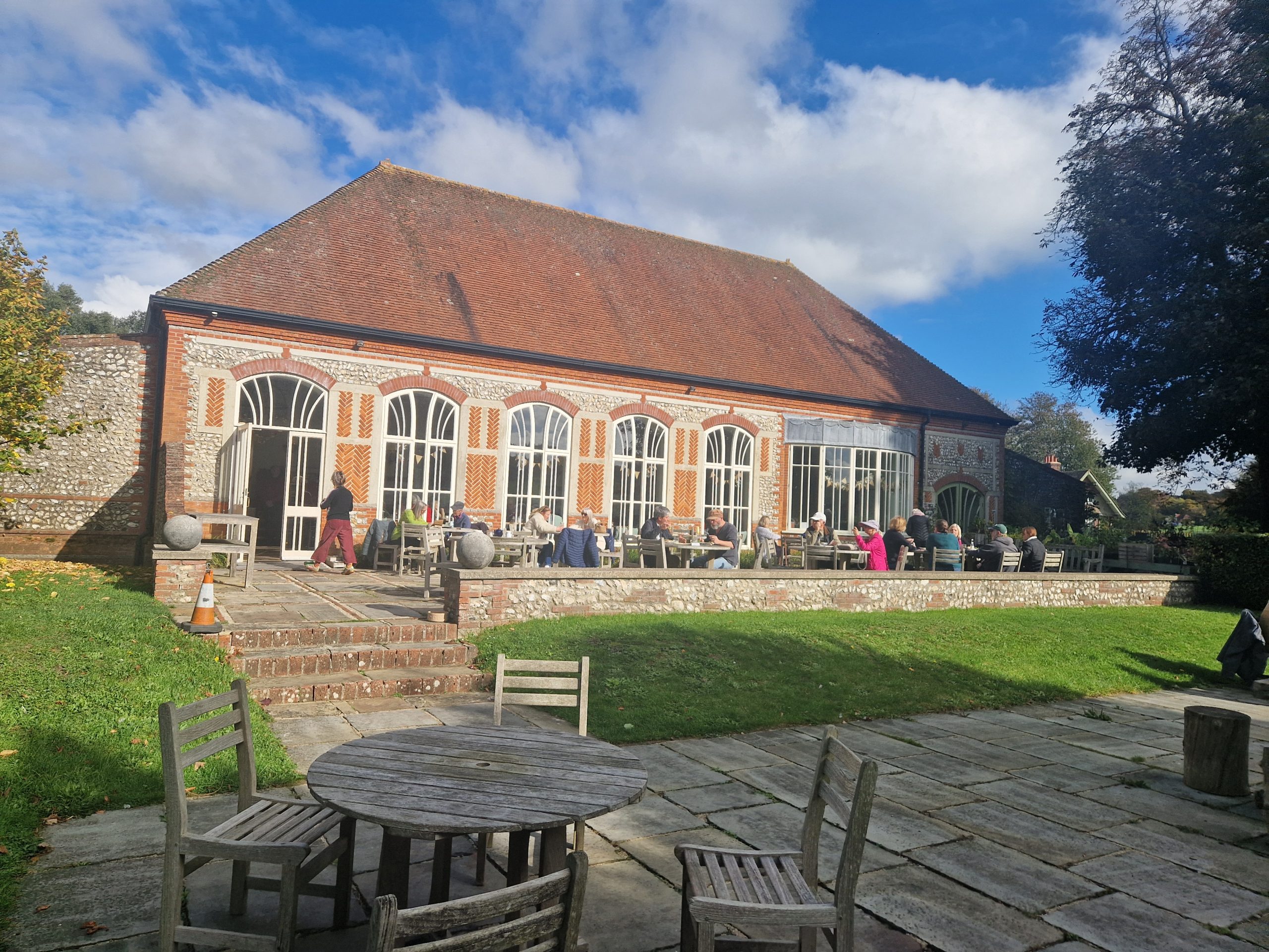 Exterior view of the West Dean Gardens Restaurant courtyard with outdoor seating, historic brick architecture, and visitors enjoying the garden setting under a blue sky.