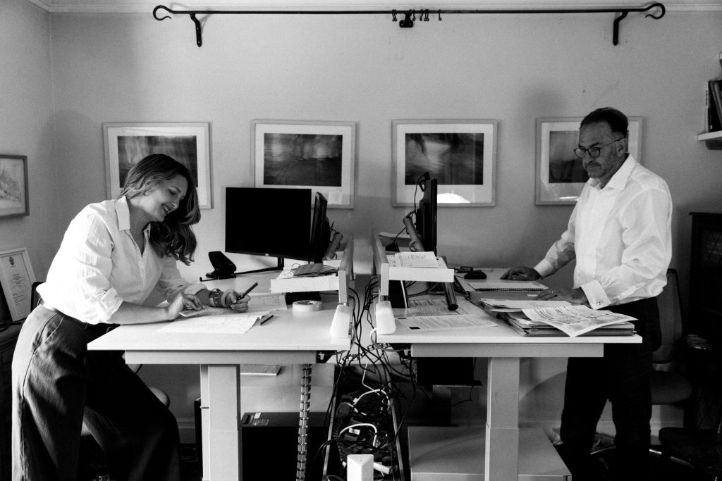 Black and white photograph of a father and daughter working opposite each other at standing desks in a small office. The daughter smiles while sketching on paper, while the father works at a computer surrounded by drawings and documents. Framed artwork hangs on the wall behind them, giving the workspace a calm, creative atmosphere.