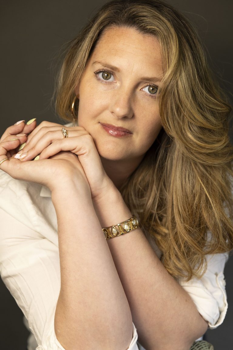 portrait of a woman with long wavy hair resting her hands gently beneath her chin while looking toward the camera. Soft lighting and a relaxed pose create a thoughtful, approachable, and creative atmosphere.