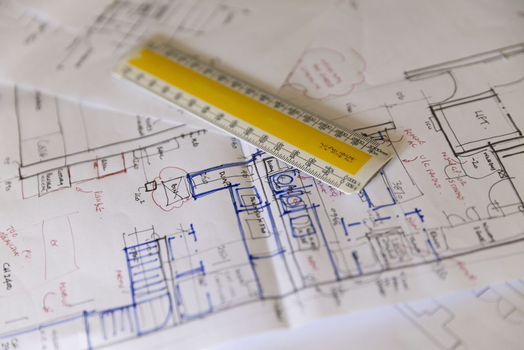 Close-up photograph of architectural and foodservice design drawings spread across a desk, with a yellow ruler placed across the plans. The image highlights the technical planning and spatial coordination involved in hospitality and commercial kitchen design.