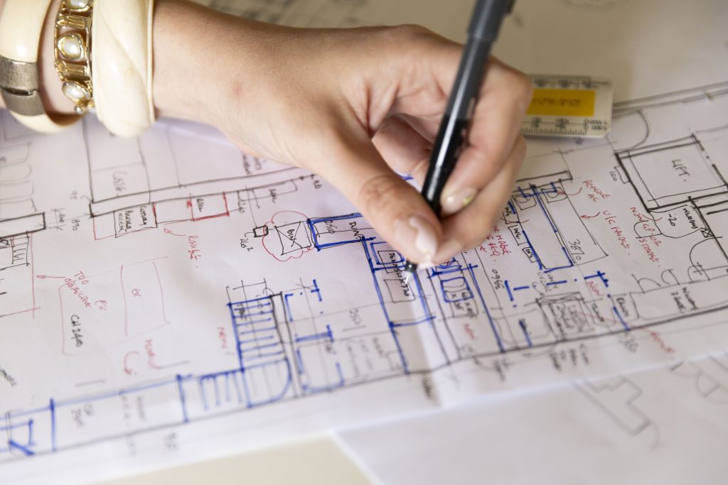 Close-up photograph of a hand sketching technical layout drawings on paper with a pen. The image focuses on the design process, showing detailed plans and annotations spread across the workspace, conveying creativity, planning, and problem-solving.