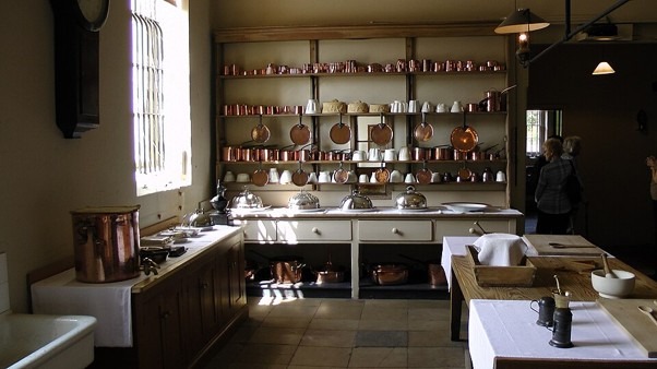 Historic kitchen at Audley End House, Essex. Long preparation tables and open shelving display copper pans, cookware and serving pieces beneath high ceilings and tall windows. The restored Victorian kitchen demonstrates the scale and organisation of food preparation in a nineteenth-century country house.