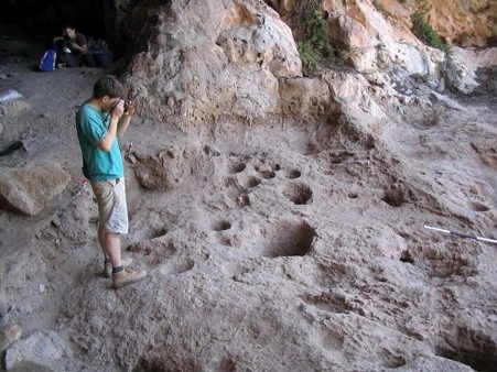 An archaeologist photographs a series of large, circular stone mortars carved into the floor of Raqefet Cave in Israel. The rock-cut hollows, varying in size and depth, were used around 13,000 years ago to process and prepare plant materials, including wild cereals and legumes, providing some of the earliest evidence of food preparation and fermentation. A rocky cave wall forms the backdrop, with another researcher seated in the shaded entrance.