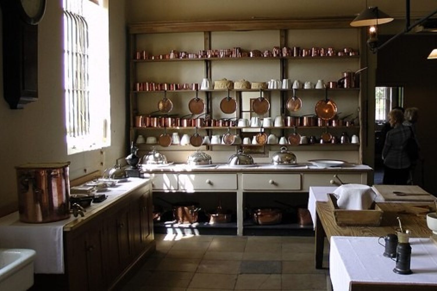 Historic kitchen at Audley End House, Essex. Long preparation tables and open shelving display copper pans, cookware and serving pieces beneath high ceilings and tall windows. The restored Victorian kitchen demonstrates the scale and organisation of food preparation in a nineteenth-century country house.