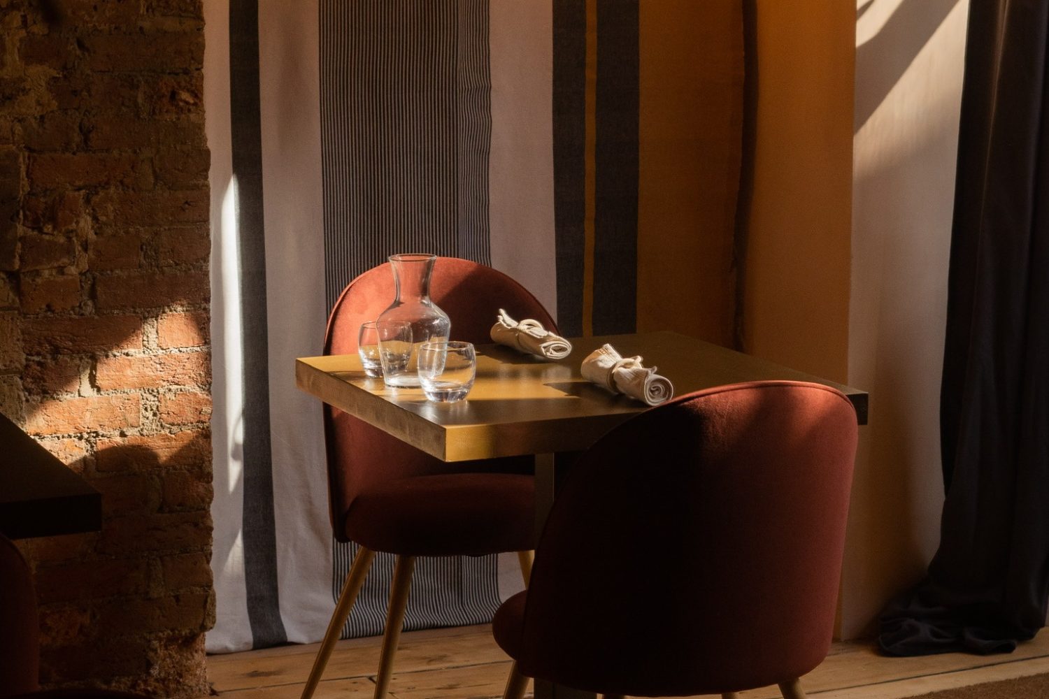 A table and chairs of a small comfortable restaurant with fabric draping walls and singular wall light. Sun casts moody shadows across scene