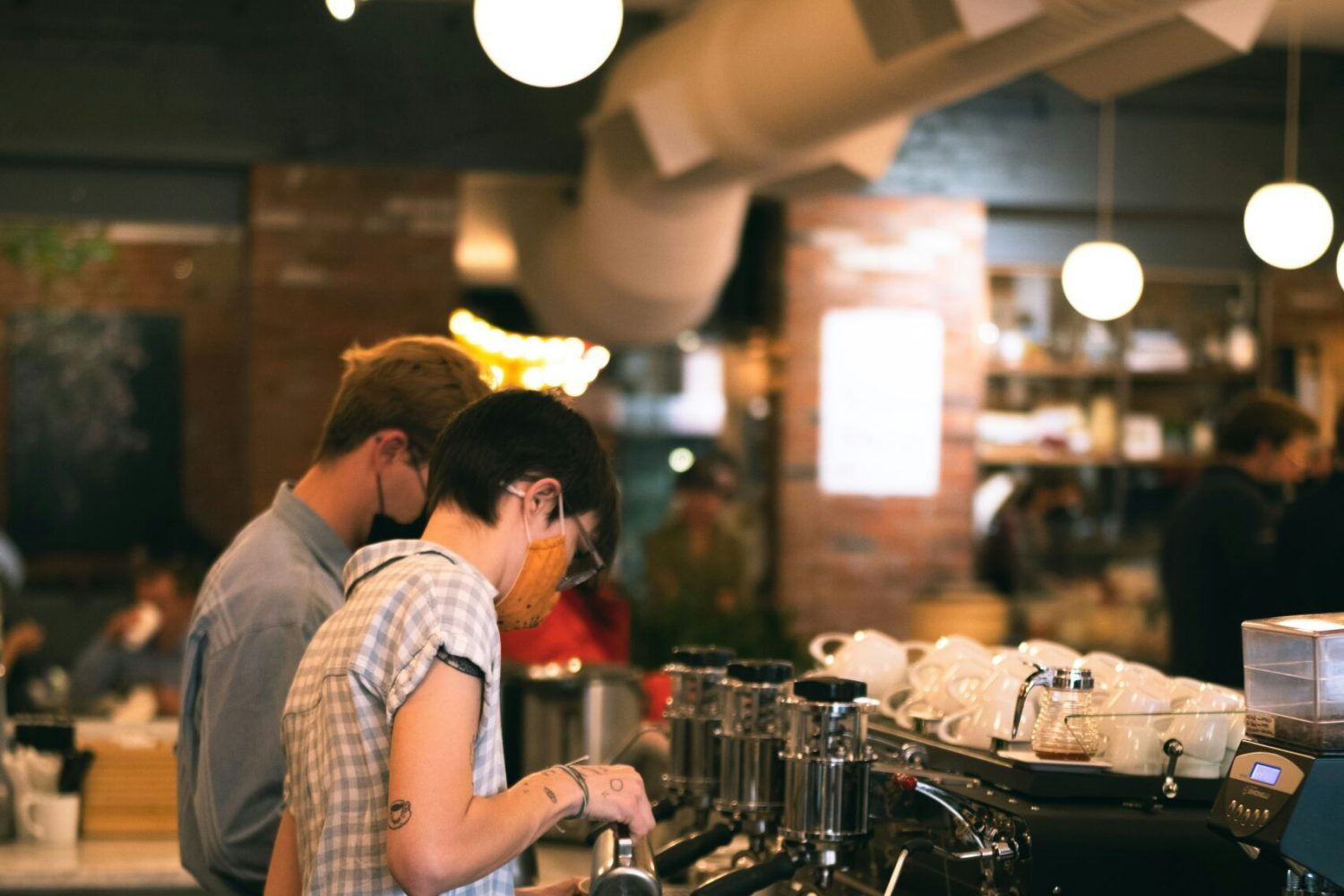 Two café staff wearing face masks working together at a 2-group coffee machine in a modern coffee shop.